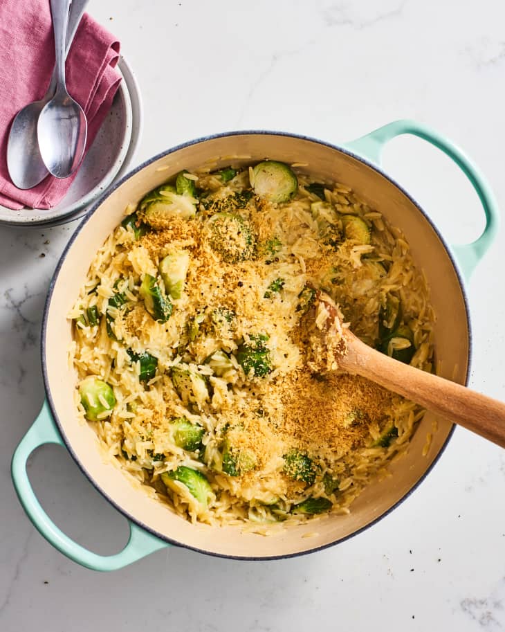 Creamy orzo with Brussels sprouts and breadcrumbs in a light blue pot, wooden spoon, and pink napkin with spoons.