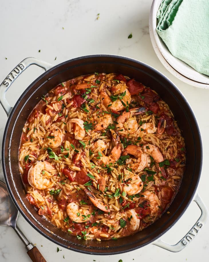 Shrimp and orzo in a black pot with tomatoes and herbs, served with a spoon.