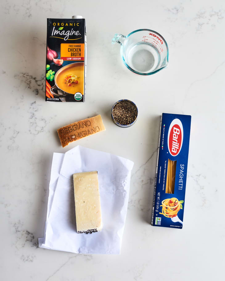 Chicken broth, spaghetti, Pecorino Romano, black pepper, and a measuring cup on a marble surface.