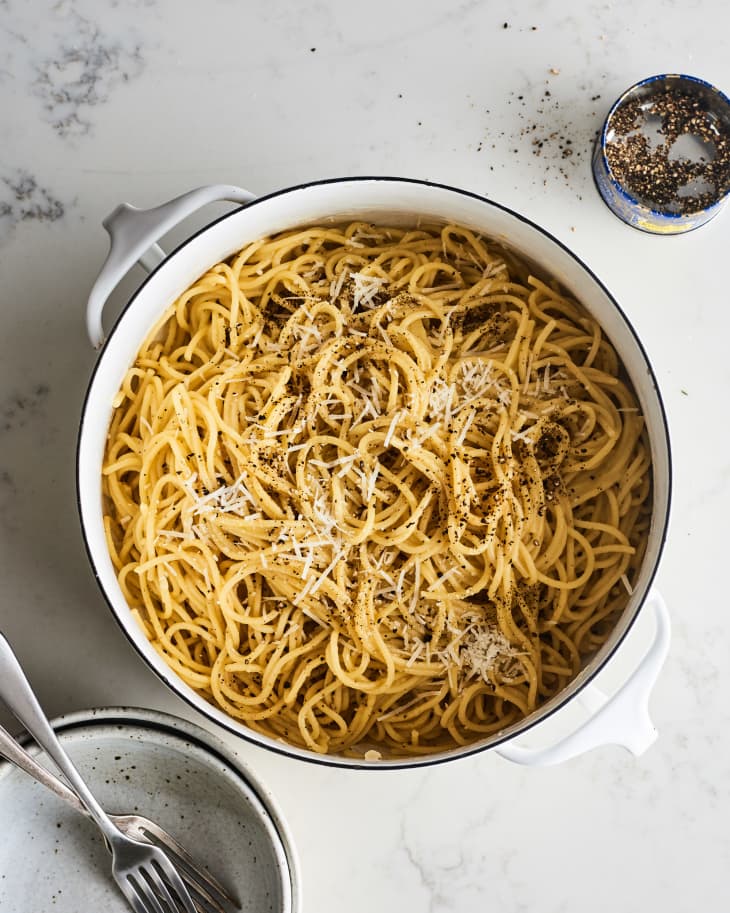 Spaghetti with grated cheese and black pepper in a white pot, next to a small bowl of pepper and a plate with forks.