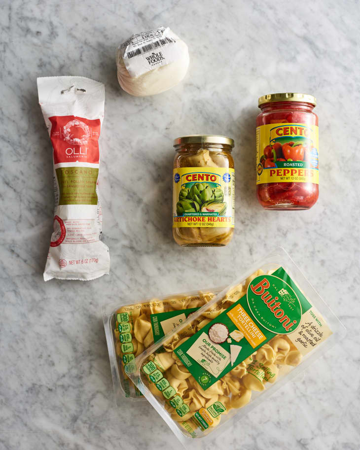 Ingredients for cheese tortellini with salami, mozzarella, artichokes, and roasted peppers on a marble surface.