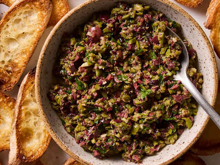 Bowl of chopped olive tapenade with a spoon, surrounded by toasted bread slices on a white plate.