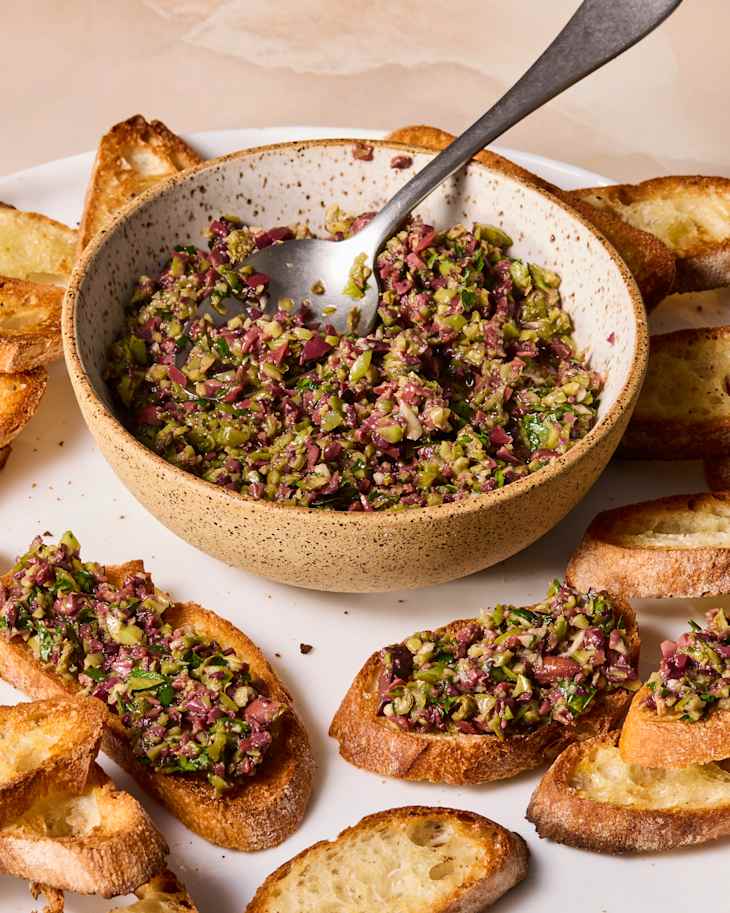Bowl of olive tapenade with a spoon, surrounded by toasted bread slices on a white plate.