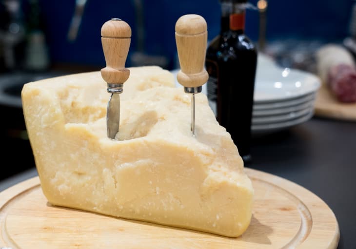Large wedge of cheese with two cheese knives on a wooden board, plates and a bottle in the background.