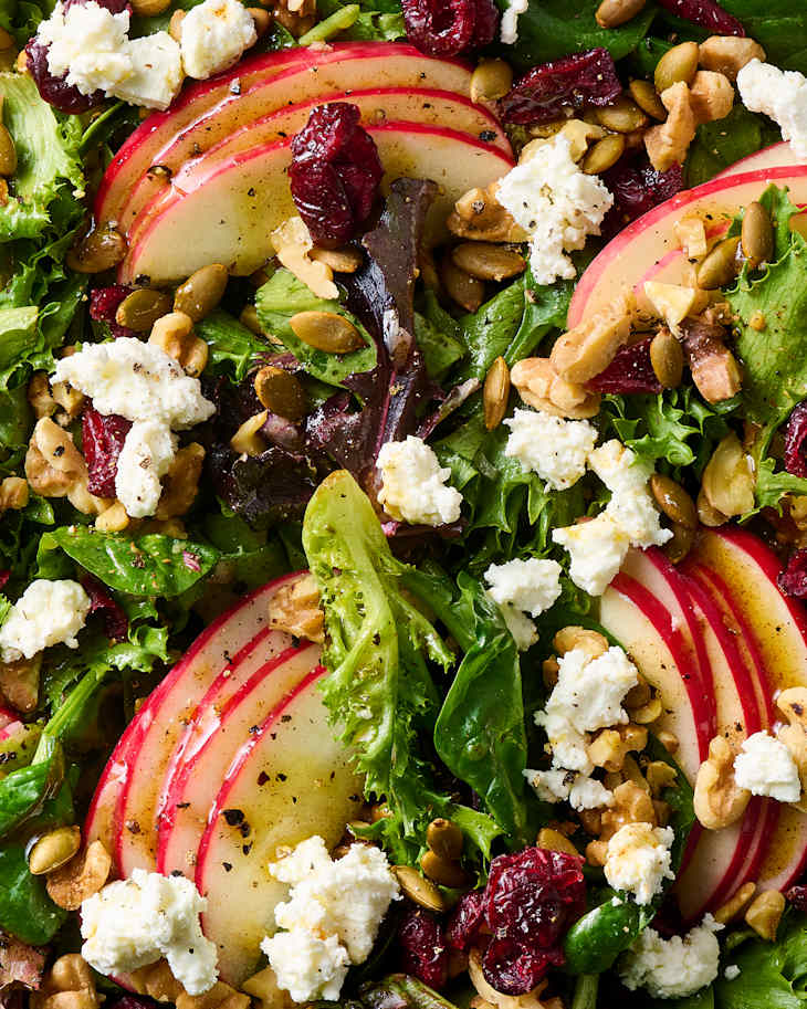 Fresh salad with mixed greens, sliced red apples, walnuts, pumpkin seeds, cranberries, and crumbled cheese.