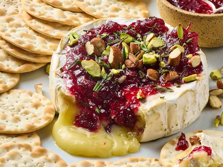 Baked brie topped with cranberry sauce, nuts, and herbs, served with round crackers on a white plate.