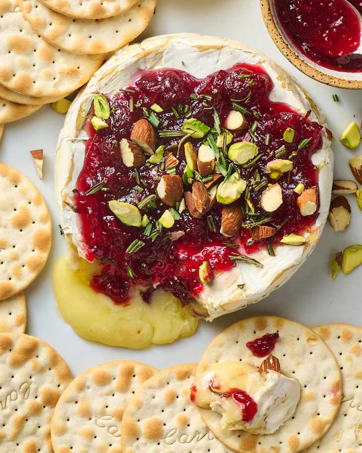 Baked brie topped with cranberry sauce, nuts, and herbs, served with round crackers on a white platter.