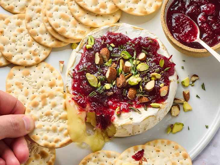 Baked brie topped with cranberry sauce, nuts, and herbs, served with round crackers on a white platter.