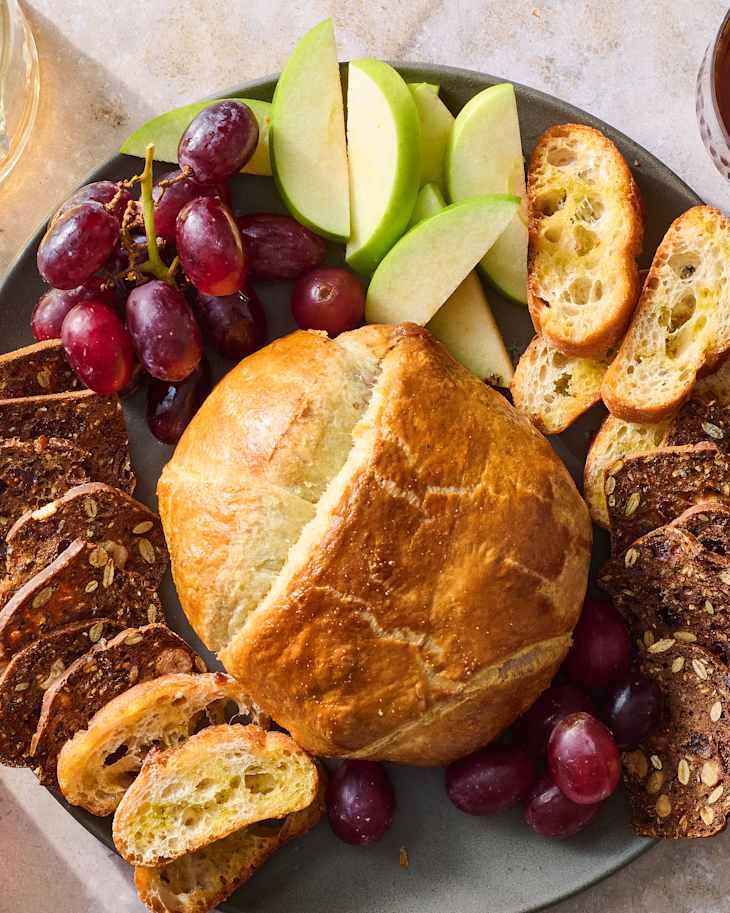 A platter featuring a brie in puff pastry, sliced apples, grapes, and toasted bread slices.