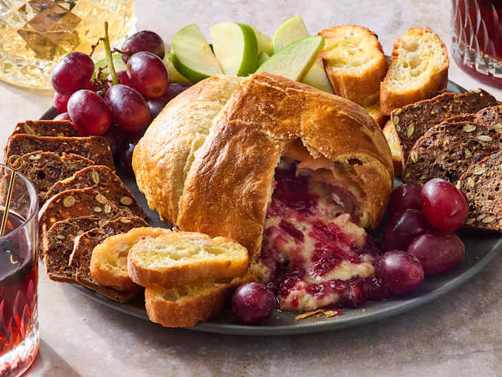 A platter featuring a brie in puff pastry, sliced apples, grapes, and toasted bread slices.