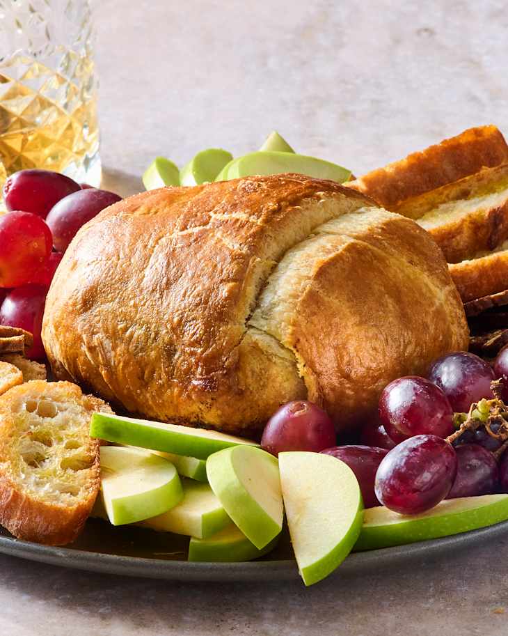 A platter featuring a brie in puff pastry, sliced apples, grapes, and toasted bread slices.
