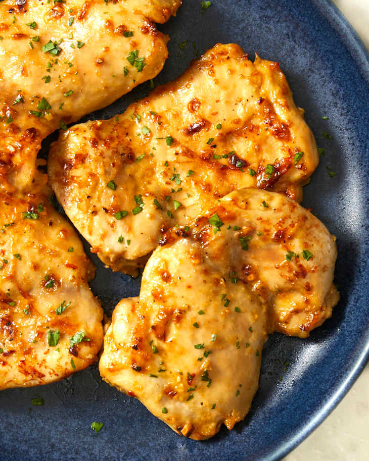 Close up of four pieces of baked boneless, skinless chicken thighs stacked on a dark blue plate on a marble background