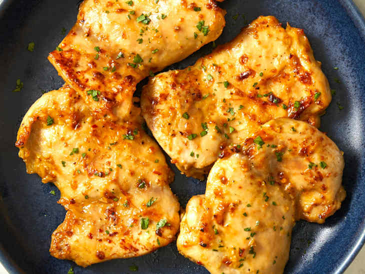 Four pieces of baked boneless, skinless chicken thighs stacked on a dark blue plate on a marble background