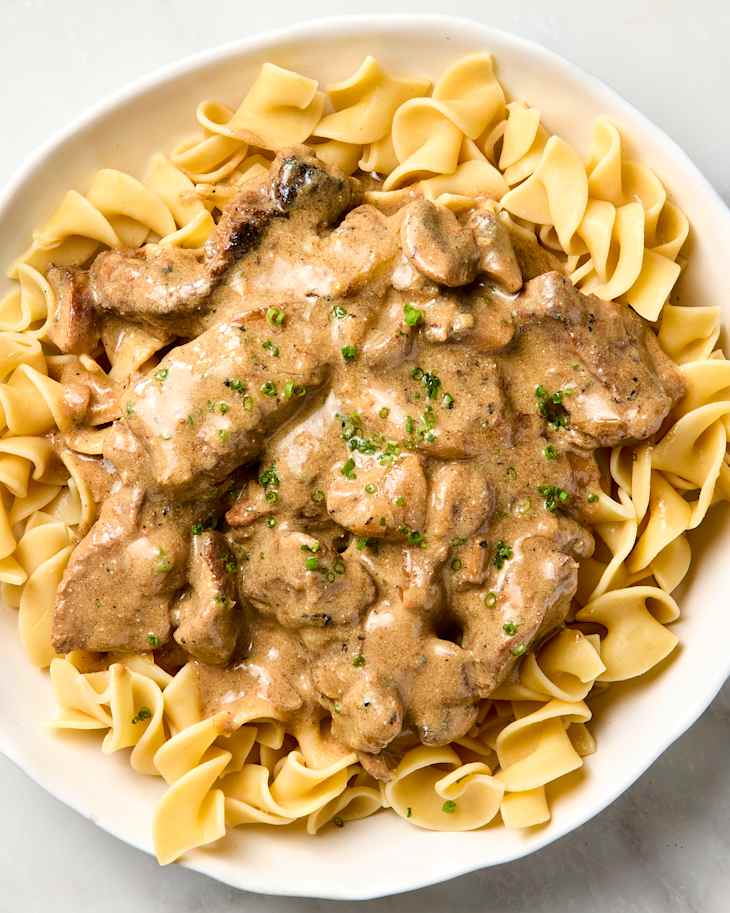 overhead shot of beef stroganoff being served over egg noodles in a white bowl.