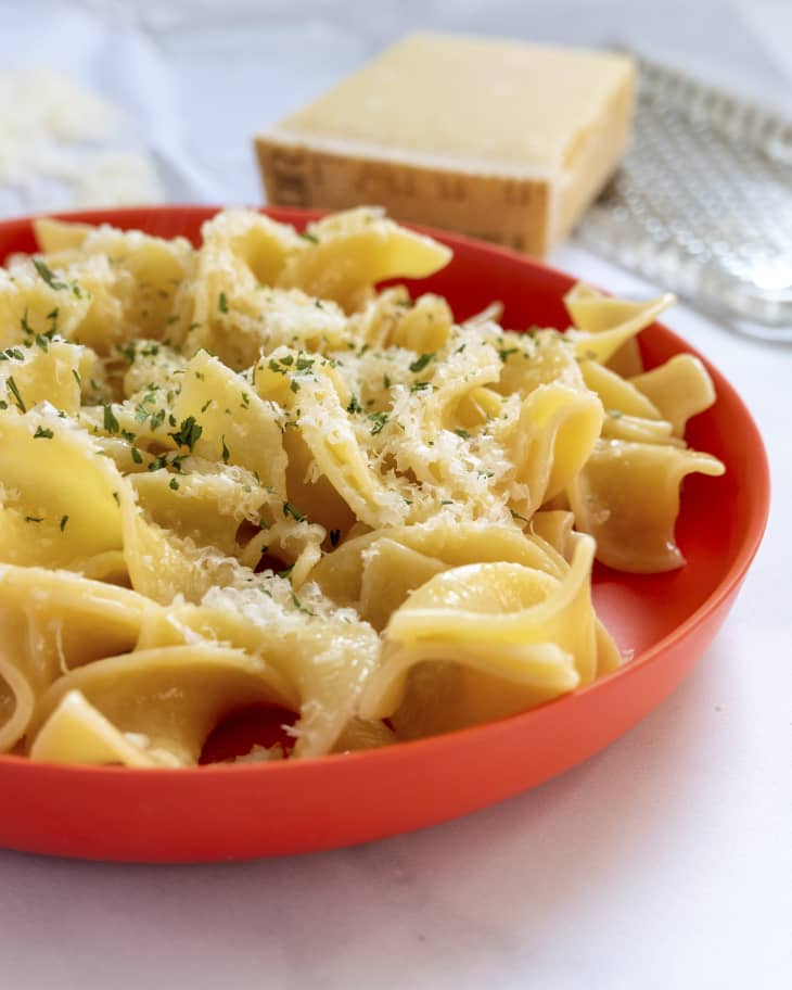 a round red plate with buttered noodles on it and a cheese grater with parmesan on the side