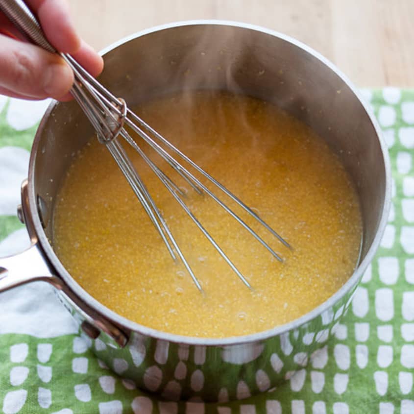 Whisking creamy polenta in a saucepan on a patterned cloth.