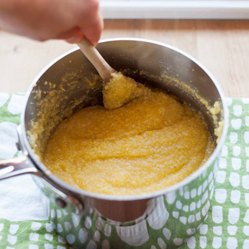 Creamy polenta being stirred in a stainless steel pot with a wooden spoon.