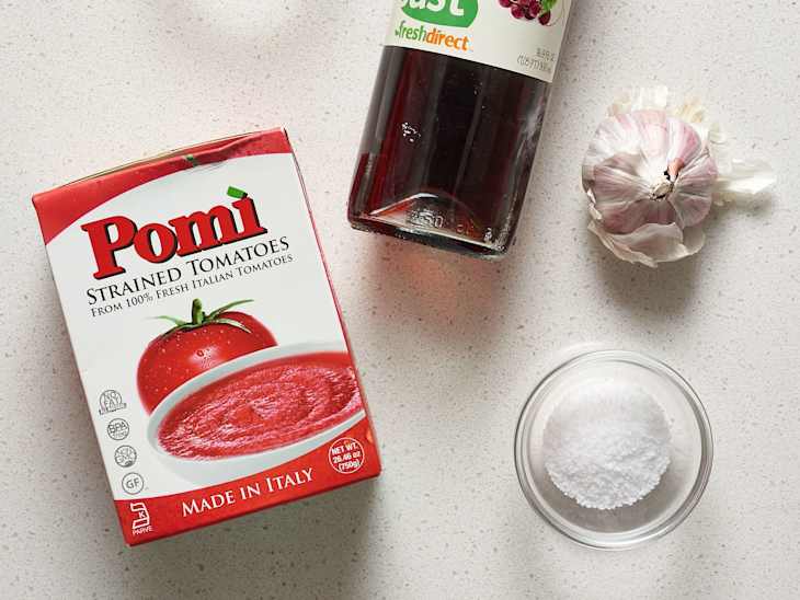 Strained tomatoes box, garlic bulb, salt in a bowl, and a bottle of vinegar on a light countertop.