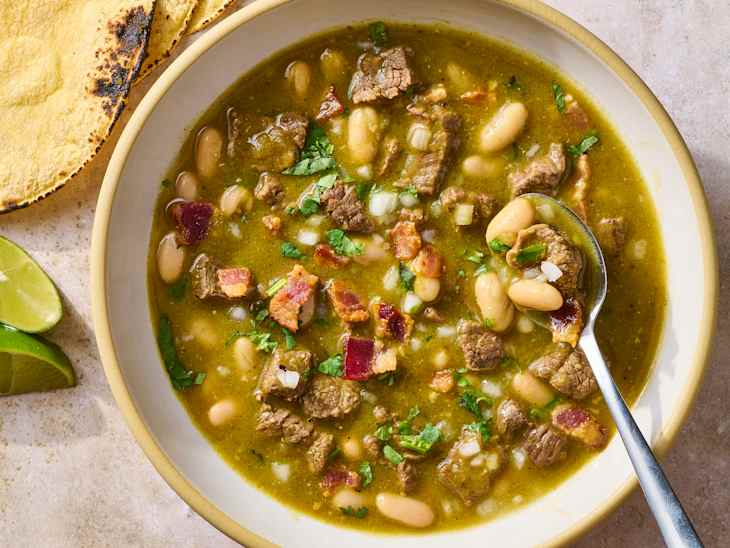 Bowl of green chili soup with beef, white beans, bacon, and cilantro, served with lime wedges and tortillas.