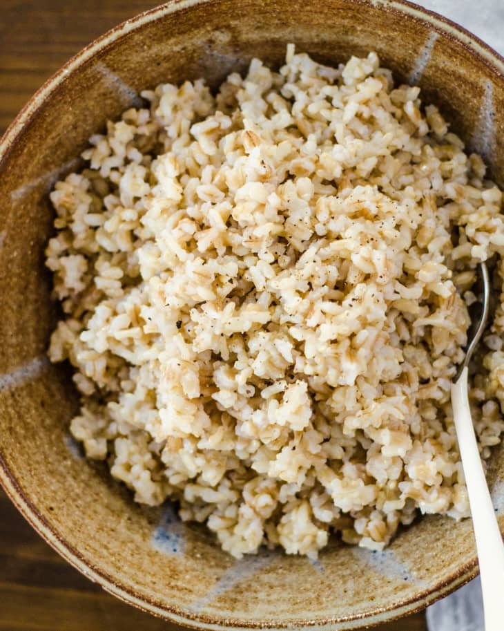 Cooked brown rice in a rustic brown bowl with a spoon on a wooden table.