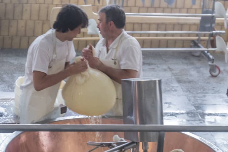 Two cheesemakers in white aprons working with a large cheese curd in a copper vat.