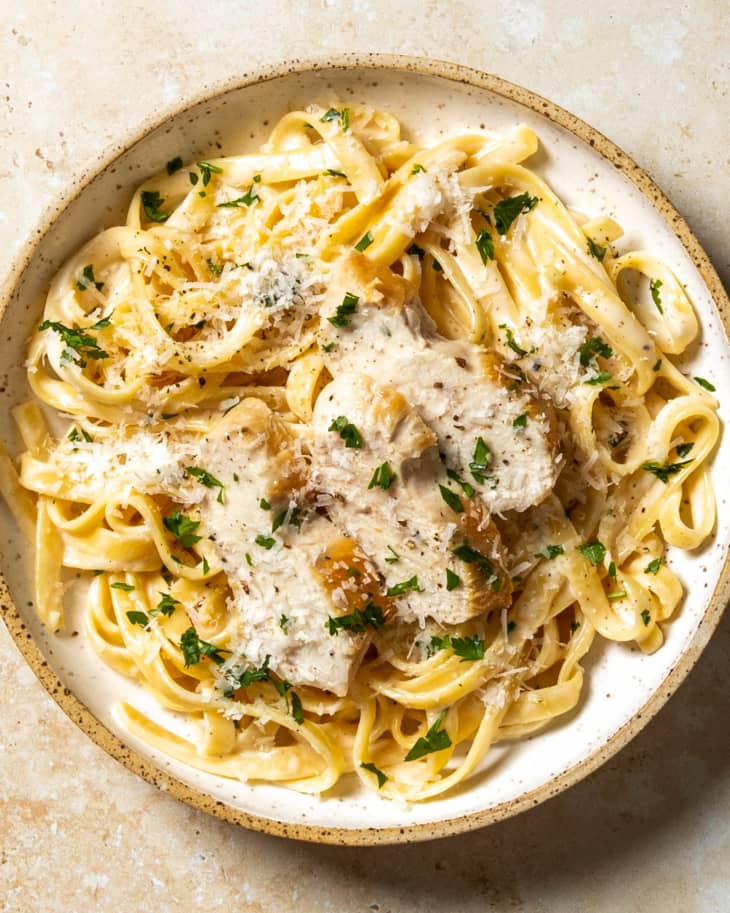 An overhead photo of a bowl of chicken fettucine Alfredo with grated parmesan cheese and parsley on top on a tan stone background