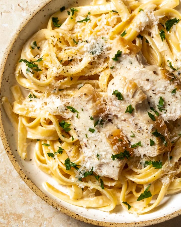 An overhead photo of a bowl of chicken fettucine Alfredo with grated parmesan cheese and parsley on top on a tan stone background