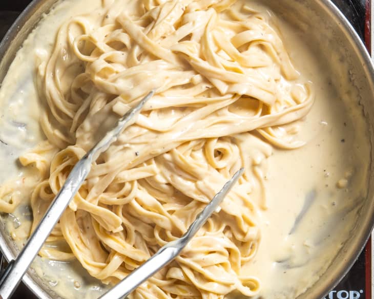 Pasta added to creamy alfredo sauce in a skillet, getting tossed with silver tongs