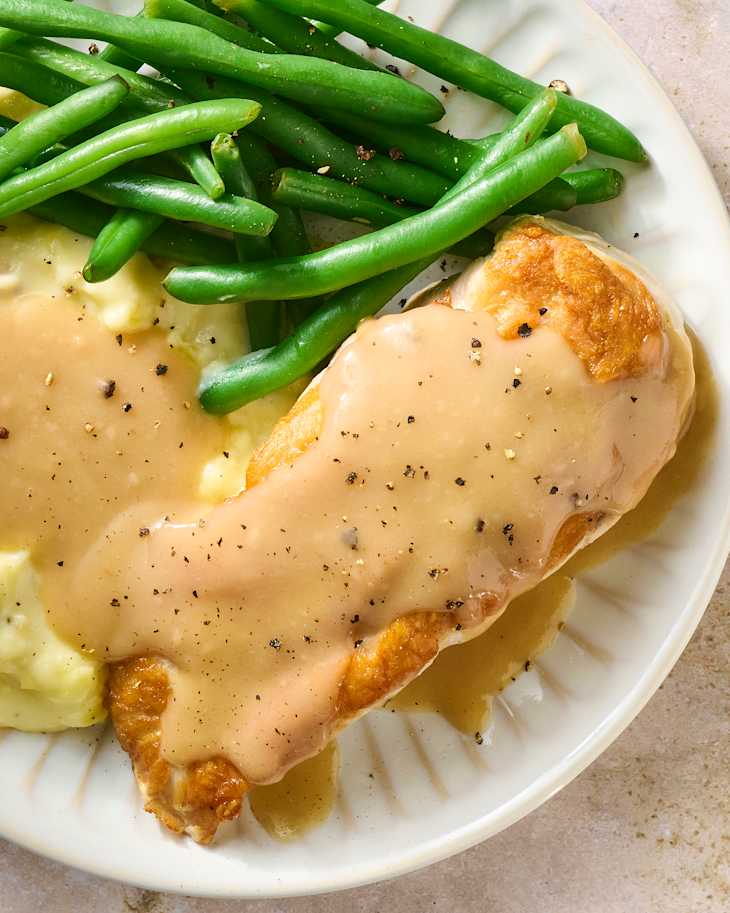 Plate of chicken smothered in gravy, served with mashed potatoes and green beans.