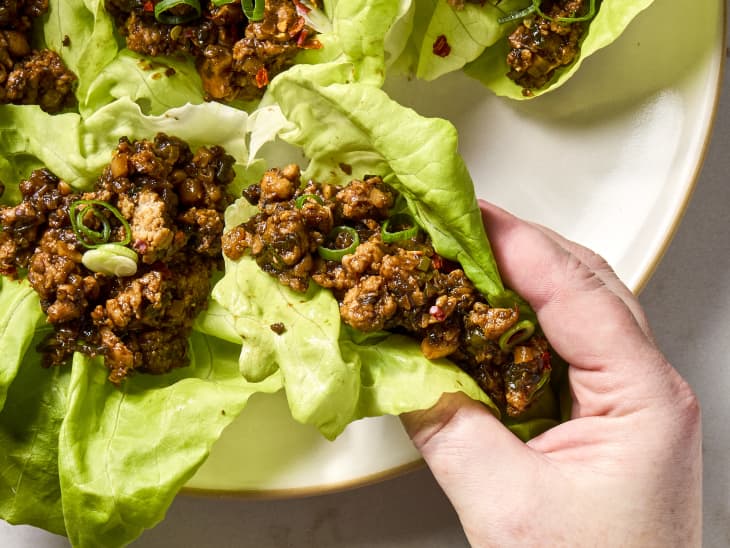 overhead shot of chicken lettuce wraps on a large white and beige trimmed platter, with a hand coming in and grabbing a wrap
