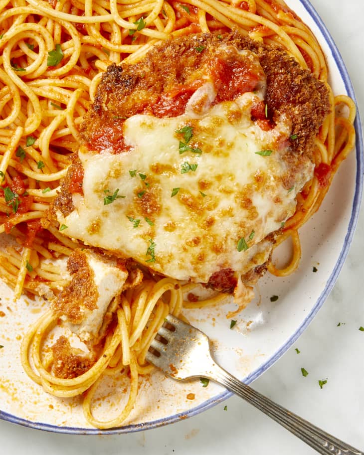 Overhead view of chicken parmesan over spaghetti on a white plate with a blue outer rim, and a fork resting on the plate.