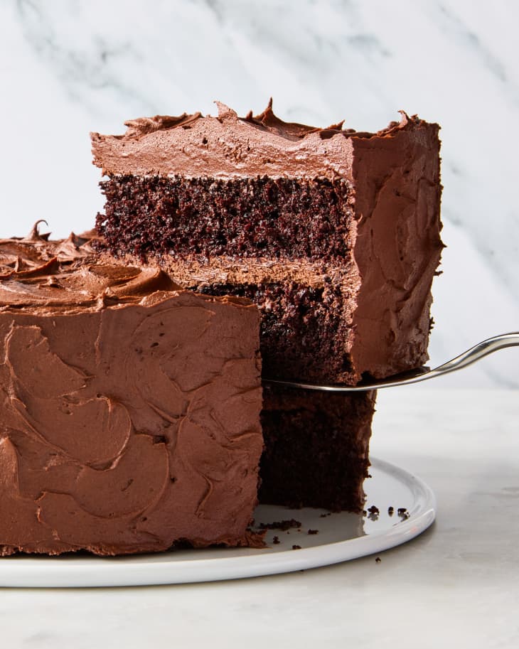 head on shot of a whole chocolate cake with on slice being lifted out by a silver cake cutter.