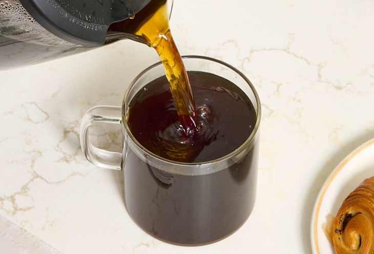 angled shot of someone pouring coffee into a glass mug, with a chocolate croissant next to the coffee