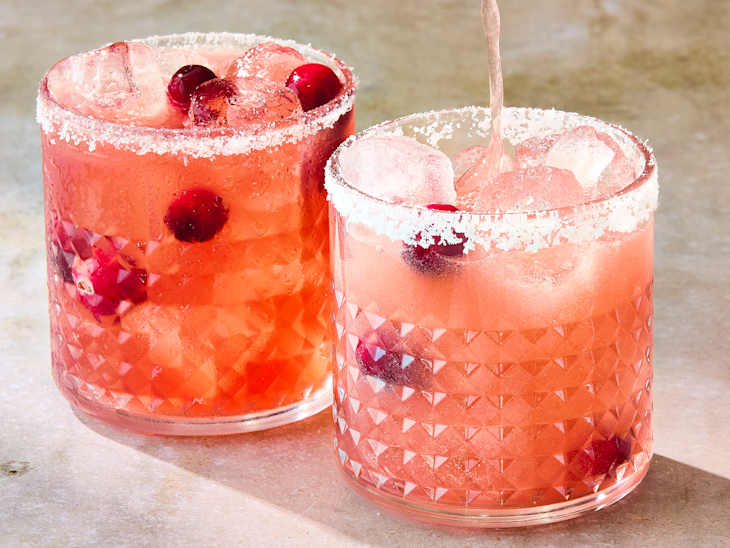 Two glasses of pink cocktails garnished with cranberries and rimmed with salt, filled with ice cubes.