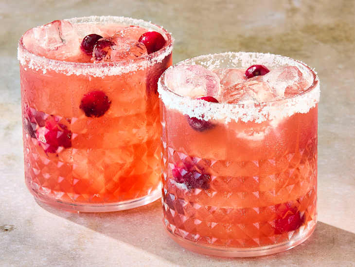 Two crystal glasses filled with a pink cocktail, garnished with ice and cranberries, rimmed with salt.