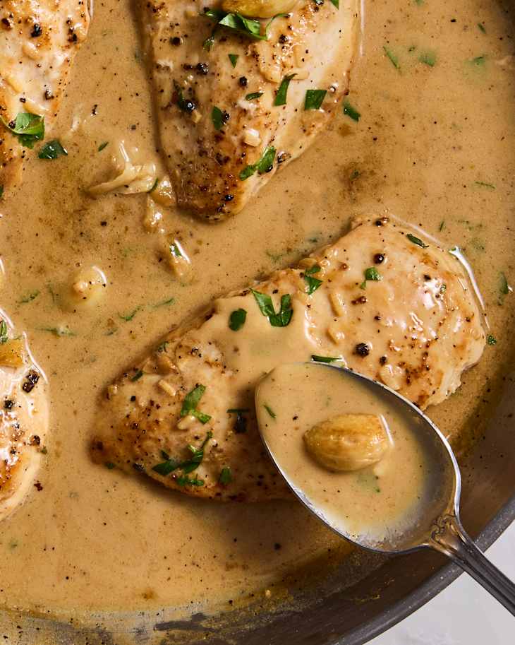 Cooked chicken breasts in a creamy sauce, garnished with parsley, and a spoonful of sauce nearby.