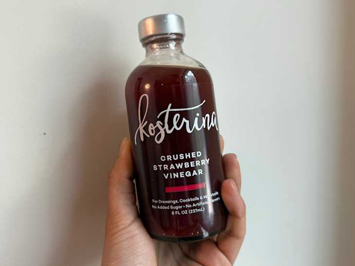 A hand holding a bottle of crushed strawberry vinegar with a silver cap and a label detailing its uses.