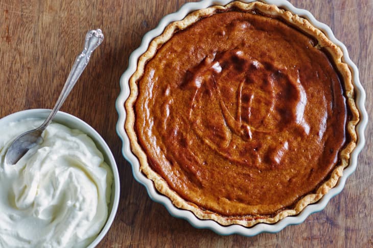 Pumpkin pie in a fluted dish next to a bowl of whipped cream with a spoon on a wooden surface.