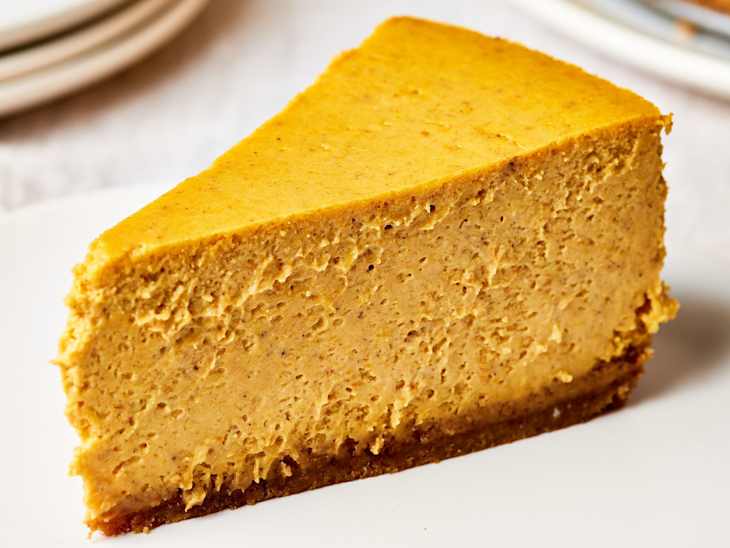 A slice of pumpkin cheesecake with the rest of the pumpkin cheesecake in the background.