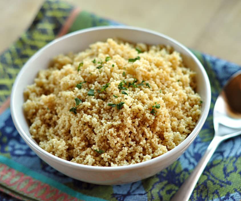 Bowl of cooked couscous garnished with herbs on a patterned cloth next to a spoon.