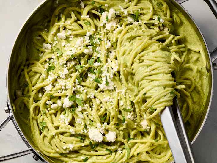 Creamy green pasta with spaghetti, topped with crumbled cheese and fresh parsley in a stainless steel pan.