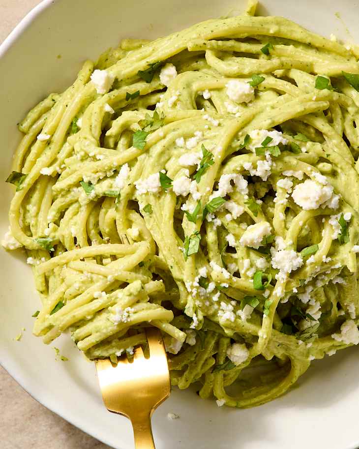 Creamy pasta with green sauce, topped with crumbled cheese and fresh herbs, served with a gold fork.