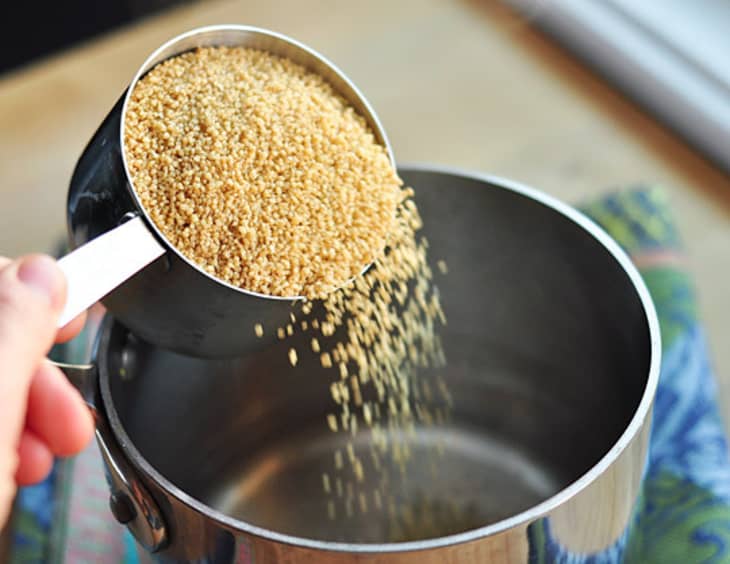Pouring couscous from a measuring cup into a stainless steel pot.