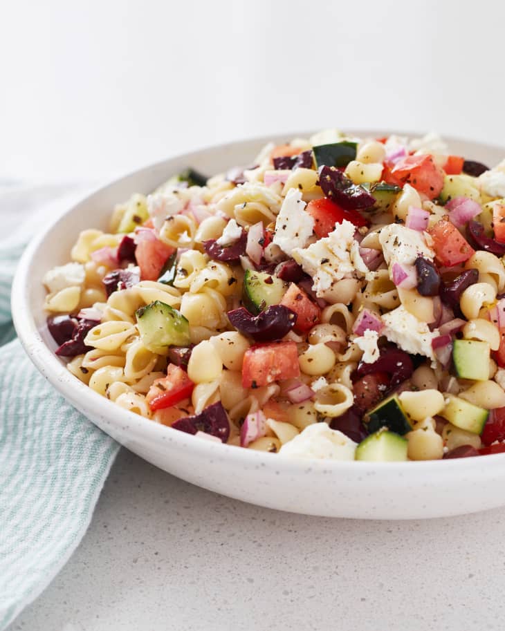 Pasta salad with cucumbers, tomatoes, olives, red onion, and feta in a white bowl on a countertop.