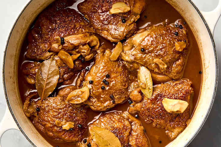 A dutch oven full of chicken thighs cooked in Filipino adobo with peppercorns and bay leaves visible.
