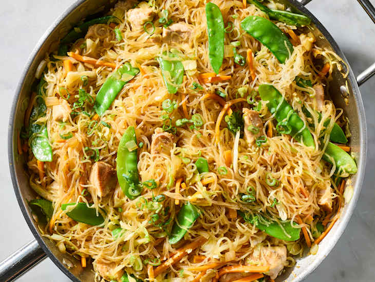 Stir-fried glass noodles with chicken, snap peas, carrots, and lime wedges in a pan