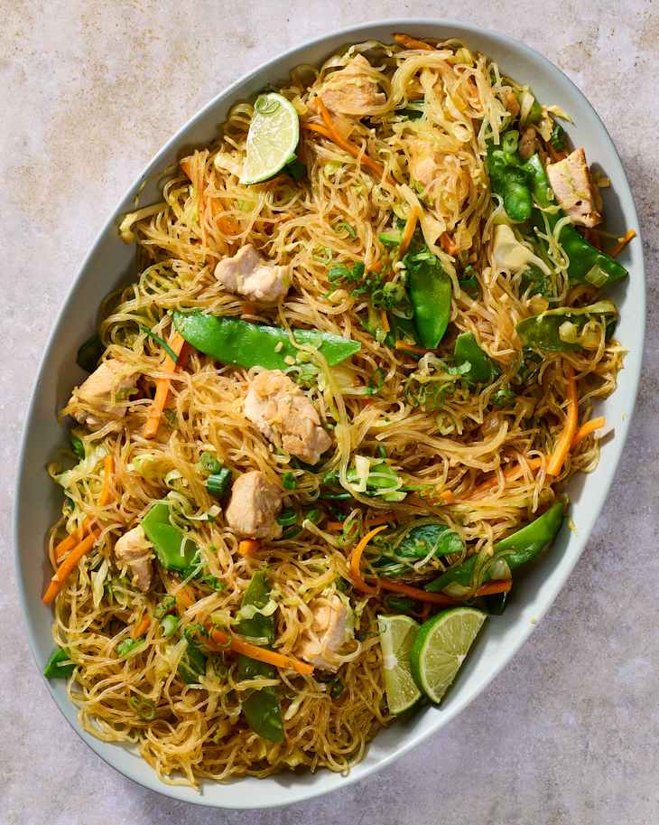 Stir-fried glass noodles with chicken, snap peas, carrots, and lime wedges in a serving dish.