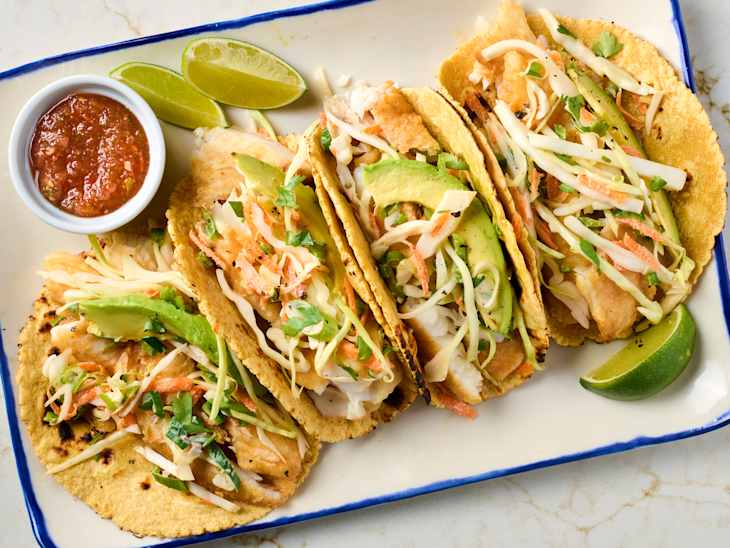 Four fish tacos topped with cabbage, avocado, and cilantro, served with a side of salsa and lime wedges.
