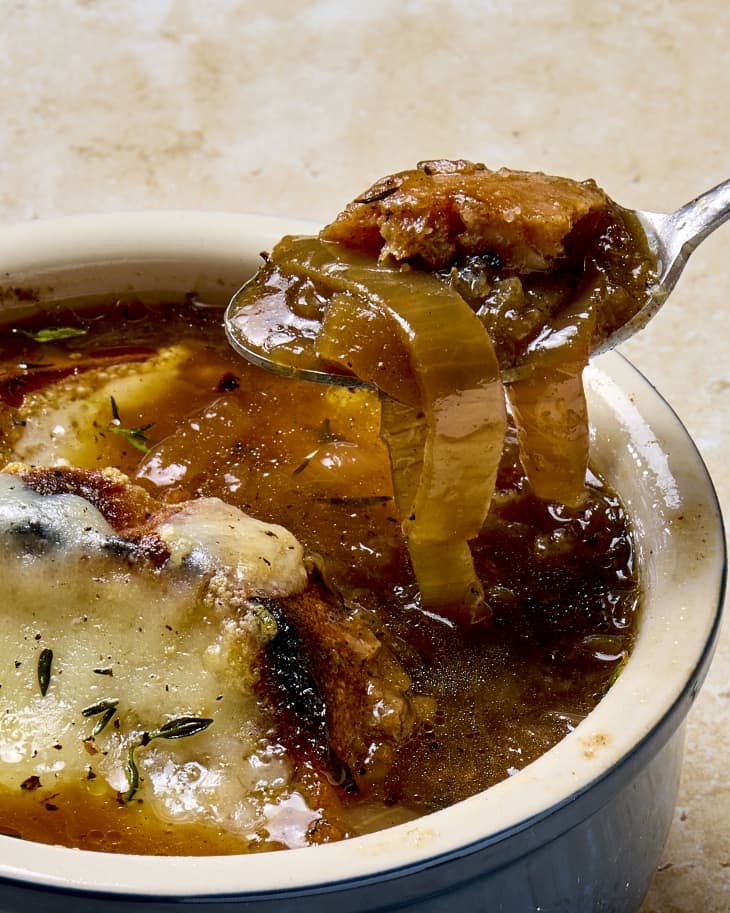 angled shot of a spoonful of french onion soup being pulled out of a navy blue soup crock.