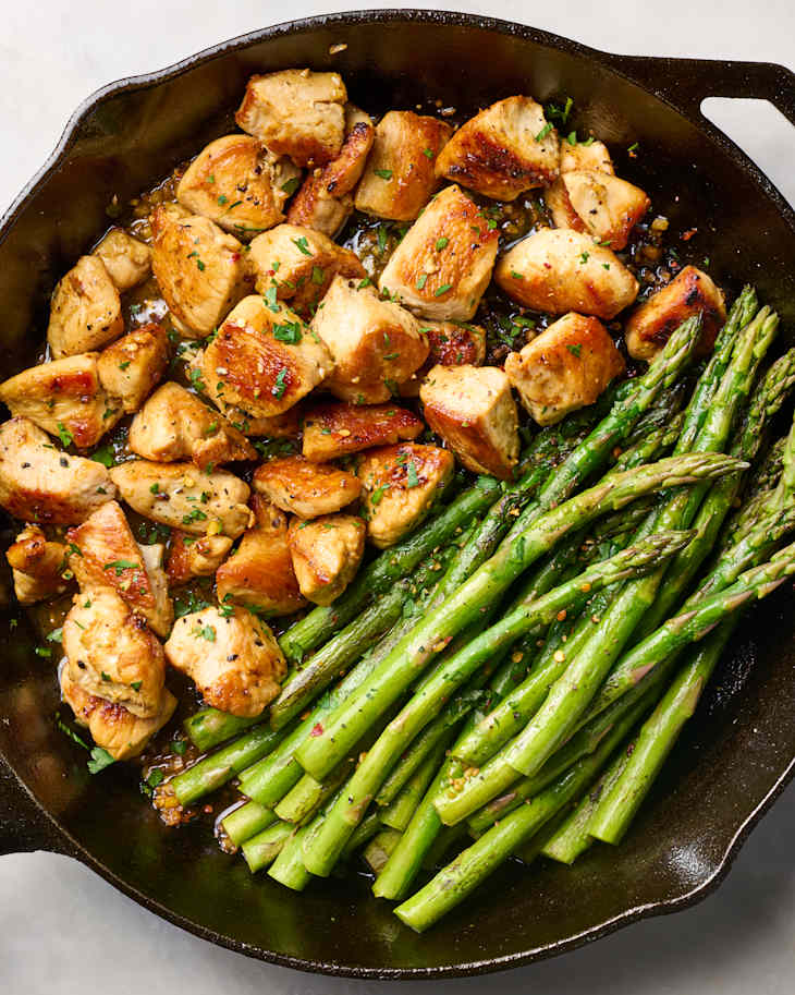 Sautéed chicken pieces with herbs alongside vibrant green asparagus in a cast iron skillet.
